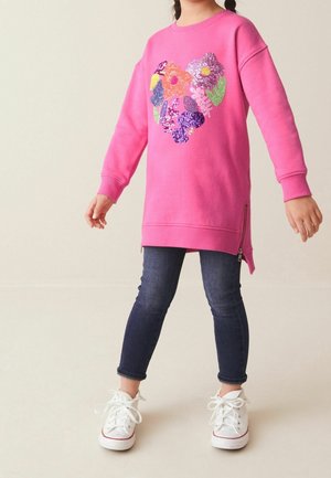 Enfant portant une robe à manches longues rose avec un motif floral coloré, un jean bleu marine et des baskets blanches, debout les bras légèrement écartés.