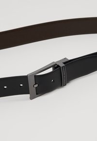 Cintura in pelle nera con una superficie liscia, dotata di una fibbia metallica rettangolare con l'etichetta "Porsche Design" in finitura opaca.