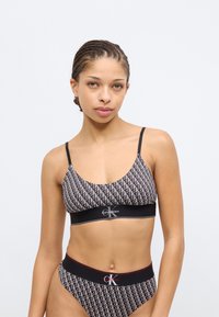 Μαύρο μπραλέτι και αντίστοιχο σλιπ με επαναλαμβανόμενο λευκό μοτίβο λογότυπου. Ρυθμιζόμενες τιράντες και ελαστική ζώνη μέσης με κόκκινες λεπτομέρειες.