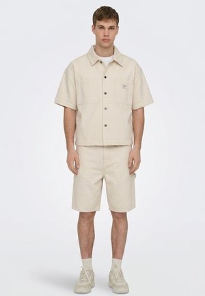 Joven de pie con una camisa color crema de manga corta abotonada, pantalones cortos a juego hasta la rodilla, calcetines y zapatillas deportivas sobre un fondo liso.