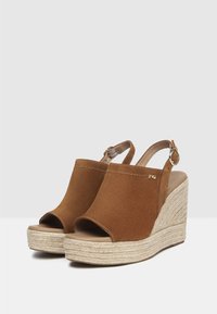 Sandales à talon compensé en daim marron avec un bout ouvert, un design à bride arrière et une semelle en corde texturée. Présente un accent en métal avec un logo sur le côté.