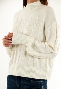 Maglione chunky bianco a cavo con colletto alto a costine, polsi a costine e orlo; presenta un piccolo accento con logo dorato vicino al fondo.