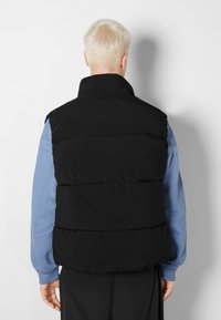 Černá prošívaná vesta s vysokým límcem, s horizontálními švy a lehkým designem, navrstvená přes modrou košili s dlouhým rukávem.