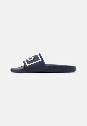 Marineblå slide-sandal med tekstureret sål og hvid omrids af firkantet og cirkulært design på den brede rem over foden.
