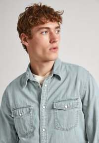 Camicia di jeans azzurro chiaro con design a bottoni, due tasche sul petto e dettagli di cucitura in contrasto. Texture morbida con una vestibilità rilassata.