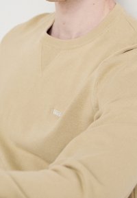 Close-up van een persoon die een beige trui met ronde hals draagt, met een klein wit logo op de borst.