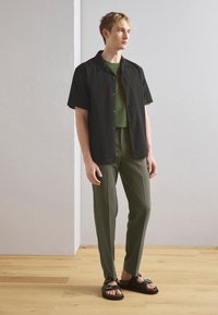 Camisa de botão de mangas curtas preta sobre uma t-shirt verde, combinada com calças verde-oliva e sandálias pretas com tiras. Fundo em piso de madeira.