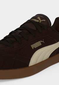 Κοντινή λήψη ενός σκούρου καφέ sneaker Puma με μαύρα κορδόνια, μπεζ πλευρική ρίγα, καουτσούκ σόλα και λογότυπο Puma στο πλάι και στη γλώσσα.