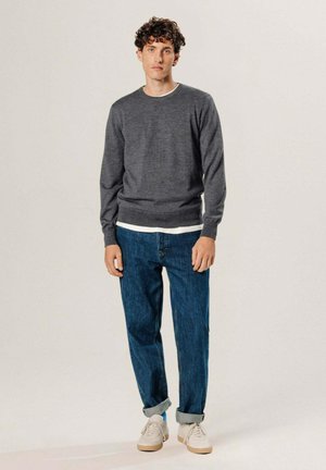 Jeune homme debout portant un sweat-shirt gris à col rond, un sous-pull blanc, un jean bleu retroussé et des baskets blanches sur un fond uni.