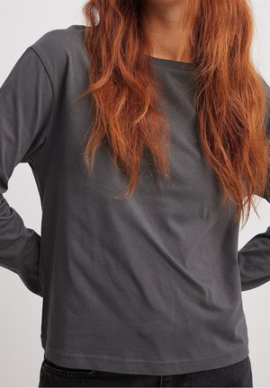 Person mit langen roten Haaren trägt ein einfaches, dunkelgraues Langarmshirt und steht mit den Händen in der Hüfte vor einem neutralen Hintergrund.