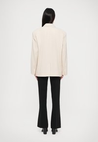 Világos bézs, oversized blazer struktúrált sziluettel, kivágott hajtókával és hátsó középen nyílással; fekete trapéz nadrágokkal párosítva.