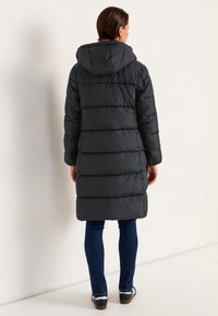 Svart puffjacka med huva, med en quiltad design, lång ärm och en nederkant som sträcker sig till mitten av låret, ihop med blå jeans och sneakers.