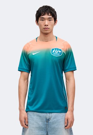 Jeune homme portant un maillot de football australien dégradé du bleu sarcelle au pêche avec le logo Nike et l'emblème national, associé à un jean bleu clair.