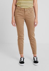 Pískové skinny džíny s hladkou texturou, pěti kapsami a zapínáním na knoflík, spárované s černými teniskami s bílými šňůrkami.