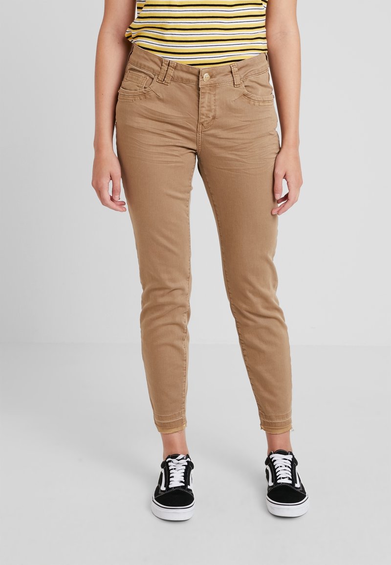 Pískové skinny džíny s hladkou texturou, pěti kapsami a zapínáním na knoflík, spárované s černými teniskami s bílými šňůrkami.