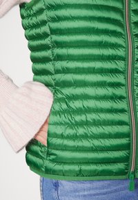 Grøn polstret vest med quiltet tekstur, der har en frontlynlås og sidelommer. Ærmet af en lyserød sweater er synligt.