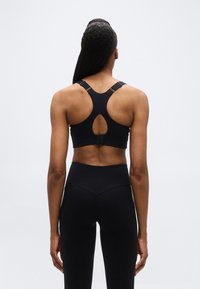 Γυναίκα που φαίνεται από π ίσω φοράει μαύρο αθλητικό σουτιέν racerback και μαύρο ψηλόμεσο κολάν απέναντι σε απλό φωτεινό φόντο.