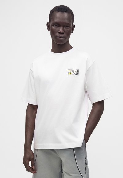 Άνδρας μοντέλο που φοράει λευκό T-shirt με γραφικά Off-White και ανοιχτό γκρι παντελόνι με μαύρο φάσα και το κείμενο Off-White στην πλαϊνή τσέπη.