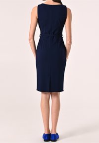 Robe sans manches bleu marine avec une coupe ajustée, longueur mi-mollet, fentes sur les côtés et une texture lisse. Associée à des chaussures en daim bleu.