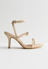 Beige högklackad sandal med flera remmar, med en knutdetalj framtill och en justerbar ankelrem. Slät, glänsande yta.