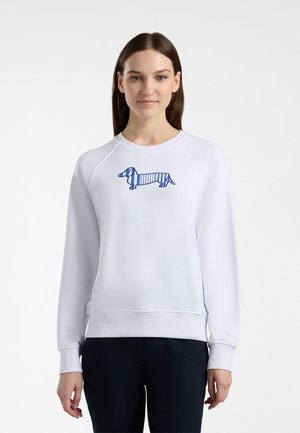 Nuori nainen, jolla on suorat ruskeat hiukset, pukeutuneena valkoiseen collegepaitaan, jossa on siniraidallinen mäyräkoira-aihe, sekä mustiin housuihin, seisomassa valkoista taustaa vasten.