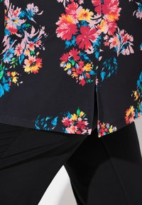 Zwarte bloes met florale print, featuring felle roze, gele en blauwe bloemen, met een gebogen zoom en zijslits. Zachte, gladde stofstructuur.