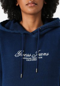Hanorac albastru marin cu șnur și capete metalice argintii. Are brodat logo-ul alb "Guess Jeans" cu textul "American Tradition California" dedesubt.