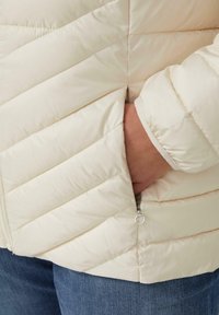 Cremfarbene Pufferjacke mit gestepptem Muster, Reißverschlusstasche und elastischen Bündchen, kombiniert mit blauen Jeans. Glattes, leichtes Material.