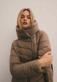 Młoda kobieta z blond włosami w taupe'owej, pikowanej kurtce puchowej z wysokim kołnierzem, z założonymi rękami, na jednolitym beżowym tle.