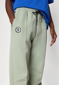 Lysegrønne joggebukser med elastisk midje, snor og svarte detaljer. Har et Chelsea Football Club-logo på låret.