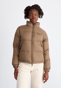 Giubbotto imbottito marrone con colletto alto, chiusura a zip, polsini elasticizzati e tasche laterali. Presenta un design trapuntato e un logo ricamato sul petto.
