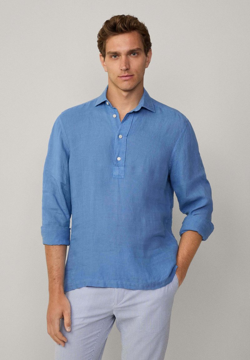 Camicia in lino azzurro chiaro con colletto, pattina dei bottoni e maniche arrotolate; abbinata a pantaloni a righe per un look casual.