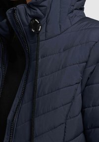 Granatowa pikowana kurtka z wysokim kołnierzem, z zapięciem na zamek błyskawiczny i regulowanym sznurkiem przy szyi. Gładka tekstura i elegancki design.