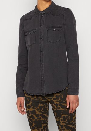 Osoba ubrana w wyblakłą czarną koszulę zapinaną na guziki z dwoma kieszeniami na piersi oraz spodnie w brązowy wzór w cętki, stojąca z rozluźnionymi ramionami.