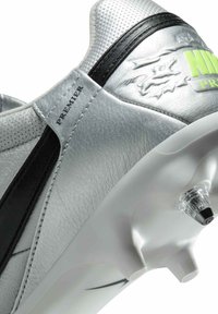 Chaussure de soccer en cuir argenté avec des accents noirs et une texture perforée. Présente un marquage vert et un point en plastique transparent sur la semelle.