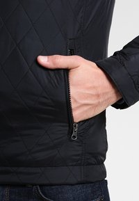Sort quiltet jakke med diagonale diamantmønstre, en lynlåslomme på siden og en glat, vandafvisende tekstur.