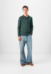 Tummanvihreä pitkähihaine polo-paita, johon on yhdistetty vaaleansiniset leveälahkeiset farkut ja ruskeat kengät, yksinkertaista valkoista taustaa vasten.