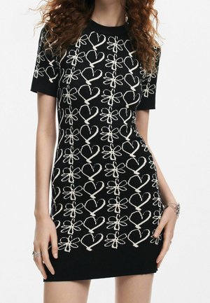 Femme portant une robe noire à manches courtes ornée de motifs de cœurs et de fleurs blanches, accessoirisée avec des bagues et un bracelet argentés.