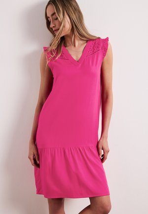 Femme portant une robe rose vif sans manches avec un col en V et un détail en dentelle, debout contre un mur clair uni, regardant vers le bas.