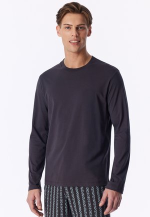 Junger Mann trägt ein schlichtes dunkles langärmeliges Shirt und gemusterte schwarz-weiße Hose und steht vor einem weißen Hintergrund.