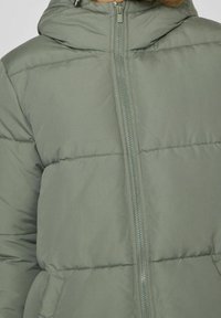 Chaqueta acolchada verde con cremallera frontal, capucha y secciones acolchadas. El material parece suave y ligeramente brillante, con bolsillos laterales.