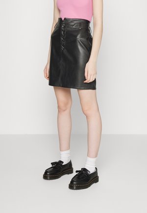 Jupe mini en cuir noir avec fermeture à boutons sur le devant, taille haute et coutures angulaires. Assortie avec des chaussures noires et des chaussettes blanches. Haut rose.