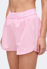 Shorts de sport Nike Pro roses avec ceinture élastique et logo Swoosh blanc portés par une personne montrée du ventre à la moitié de la cuisse.