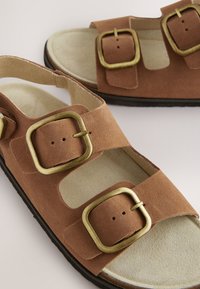 Sandalias de ante marrón con dos tiras anchas y hebillas cuadradas doradas. Plantilla texturizada y suela de goma negra. Ajustables para mayor comodidad.