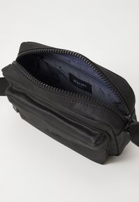 Svart tyg crossbody-väska med dragkedja upptill, som har en framficka och en märkesetikett på insidan. Texturerad yta med minimala detaljer.