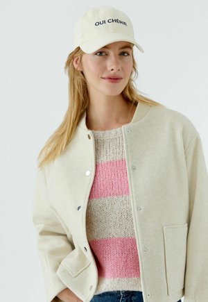 Beige Jacke aus Wollmischung mit Druckknöpfen, getragen über einem rosa gestreiften Strickpullover. Mütze mit der Stickerei "OUI CHÉRIE" in Marineblau.