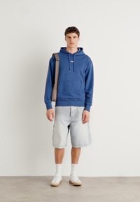 Felpa in cotone blu con tasca frontale e cordino. Pantaloni corti a righe fino al ginocchio. Calzini bianchi e sneakers. Sfondo neutro.