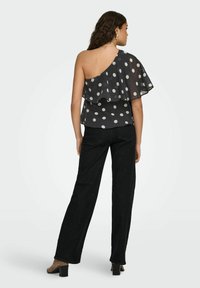 Femme portant un haut noir à pois à une épaule avec volant, un pantalon large noir et des chaussures marron à talons blocs, vue de dos.