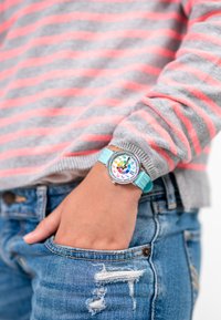 Montre colorée avec un boîtier en métal argenté, un cadran rond et un bracelet en silicone bleu clair. Présente de grands chiffres et symboles multicolores.