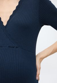 Φόρεμα σε ναυτικό μπλε με ριμπ ύφανση, βαθύ V ντεκολτέ και κυματιστά άκρα, εφαρμοστή γραμμή που αναδεικνύει τις καμπύλες και κοντά μανίκια.